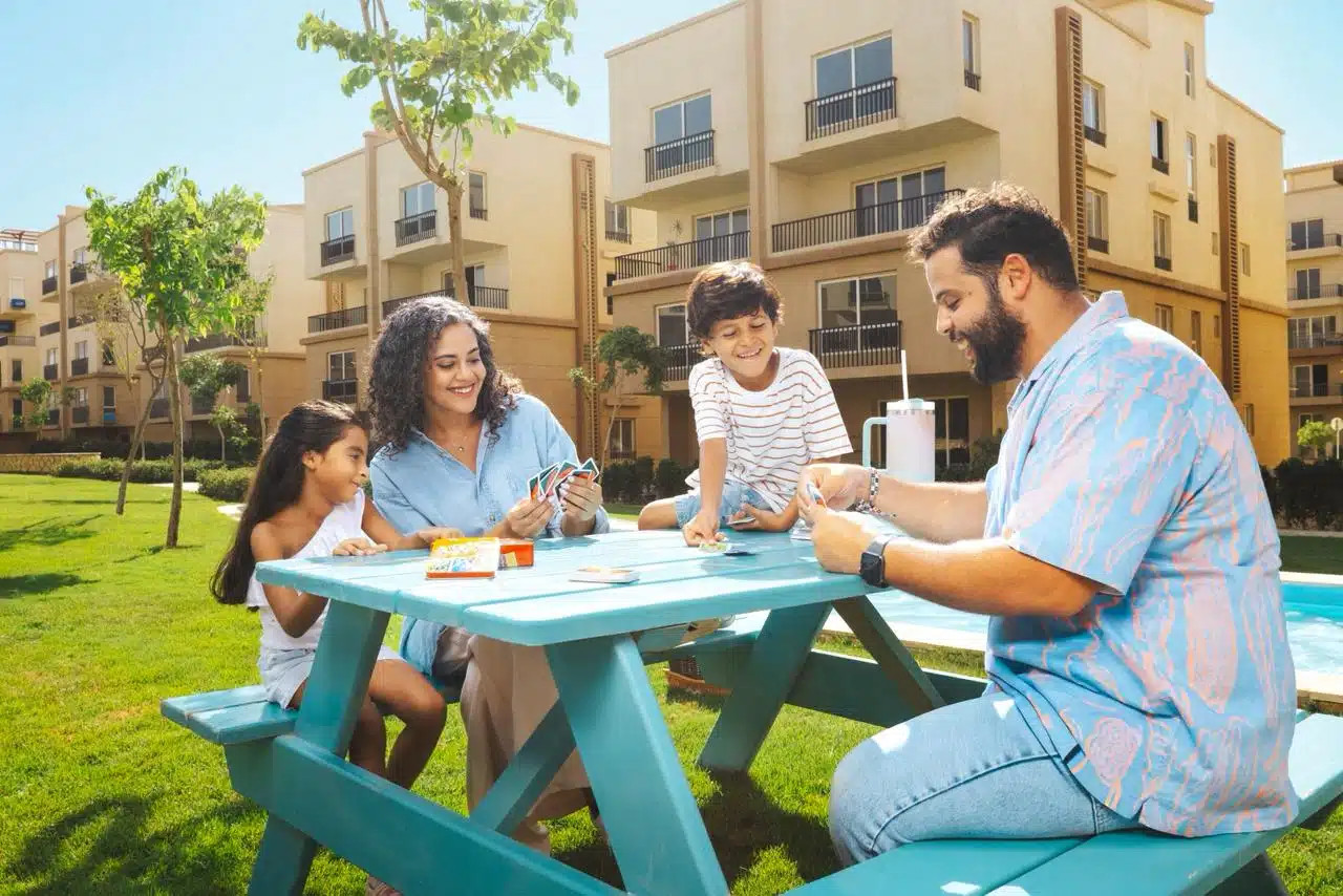 Family Of Four Playing Cards At A Picnic Table In A Sunny Courtyard With Modern Apartments And Greenery.